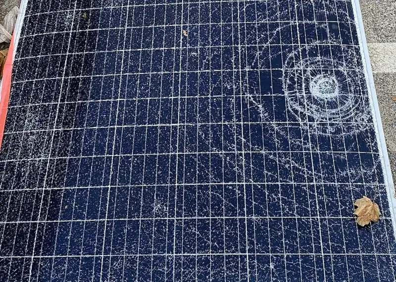 Solar module lying on pavement with shattered front glass and a ring-shaped impact point on the right