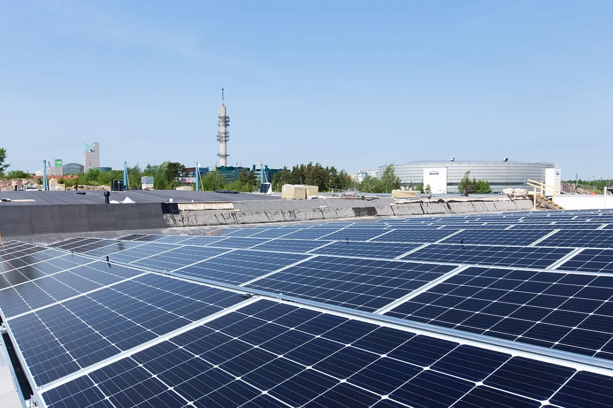 Large solar modules in rows on a flat commercial roof in Helsinki, Finland, with city buildings and a tower behind