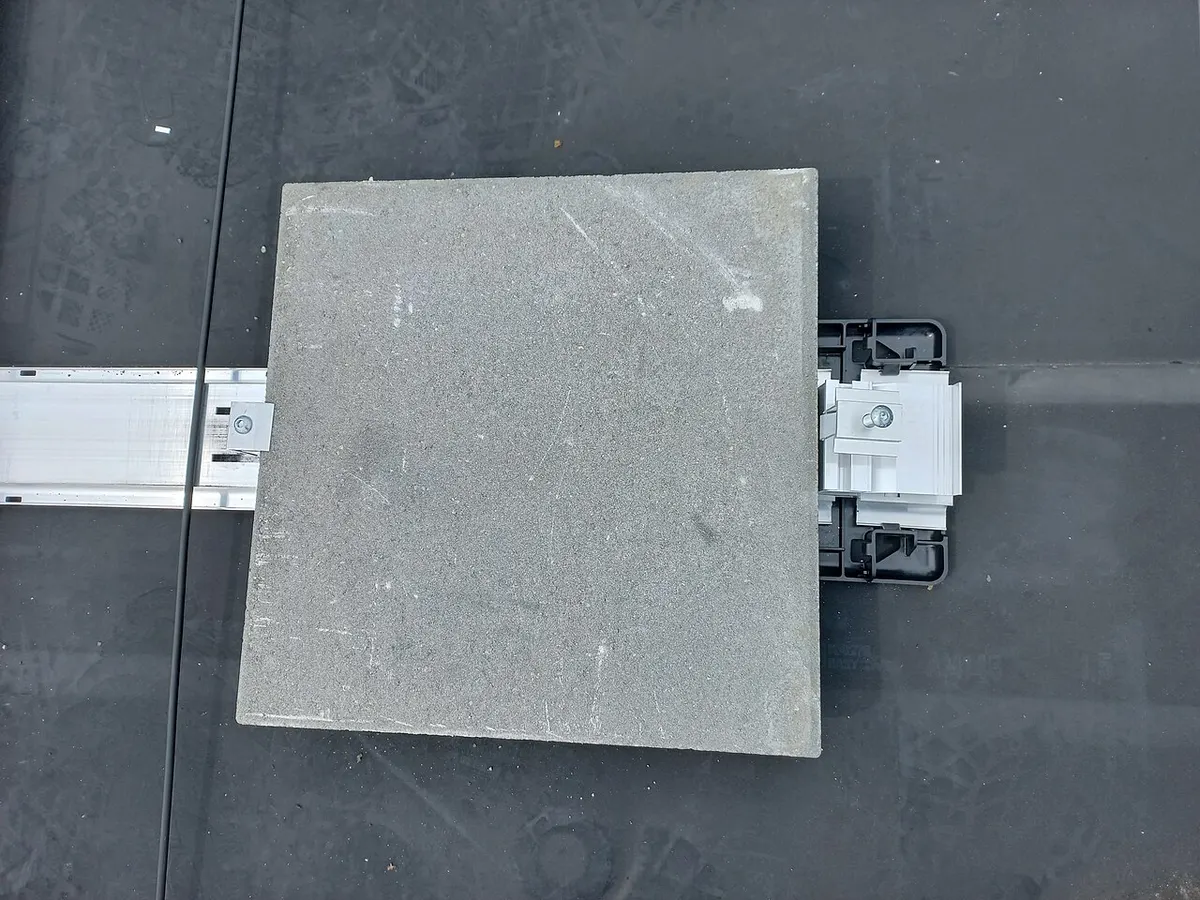 Top view of a square concrete paver laid as ballast on an aluminium mounting rail and plastic foot on a black flat-roof membrane