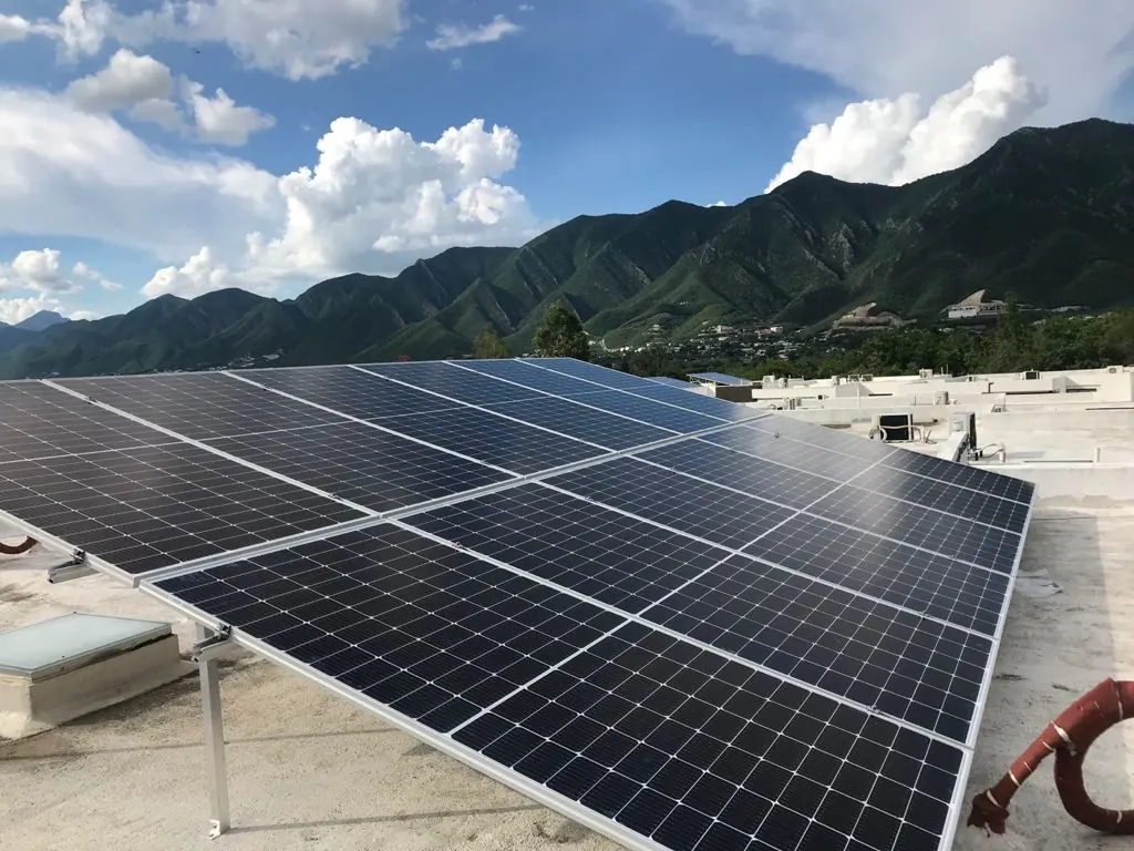 Rows of dark solar modules on tilted mounts on a flat roof with mountains in the background