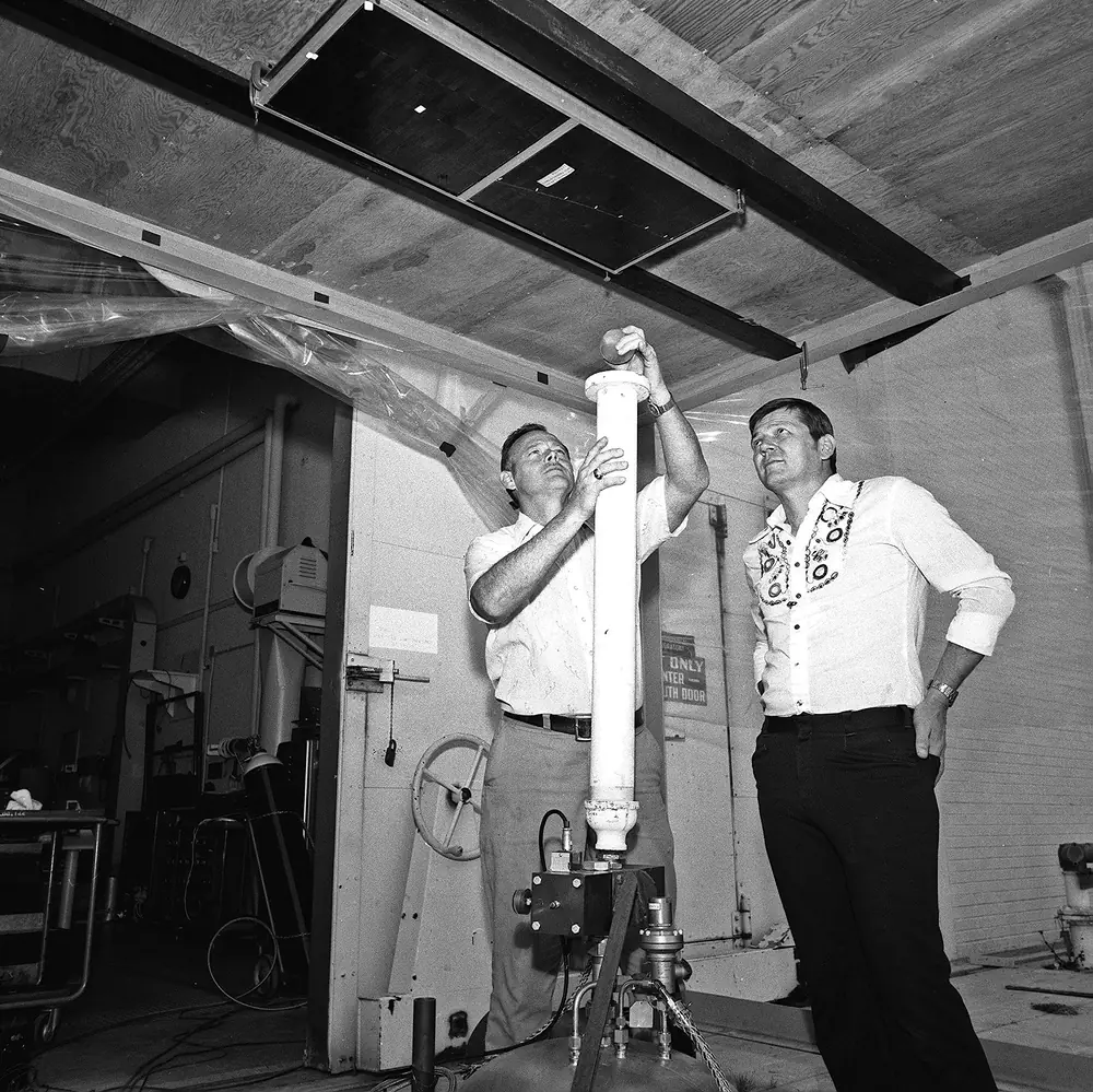 Two engineers in a 1979 test hall loading an ice ball into a vertical white launcher barrel aimed at a solar panel fixed to the ceiling above them
