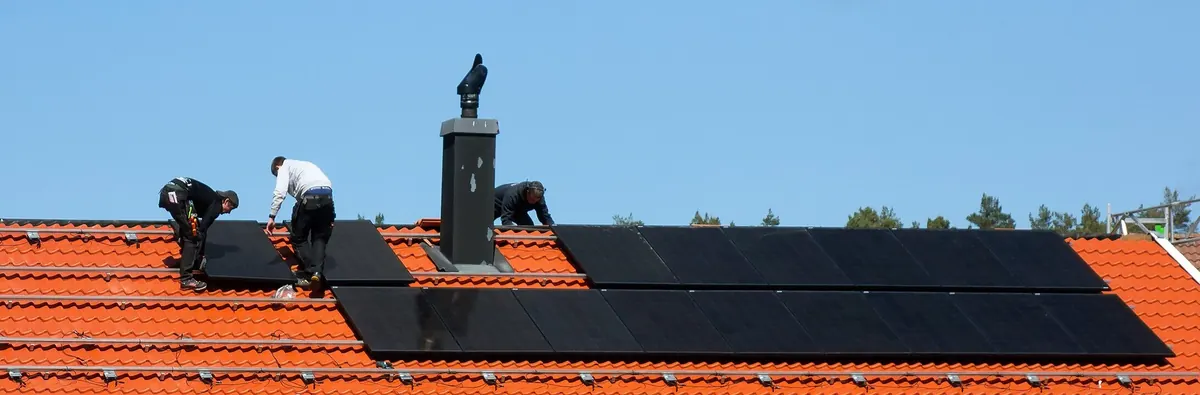 Three installers on a tiled house roof fitting black solar modules in rows around a chimney