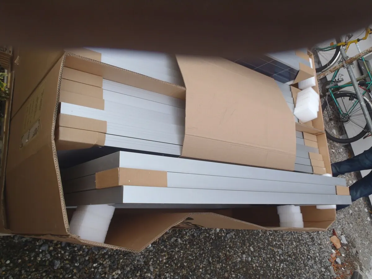 Open cardboard cartons holding solar modules packed edge-on with corner protectors and foam blocks, standing on gravel outdoors