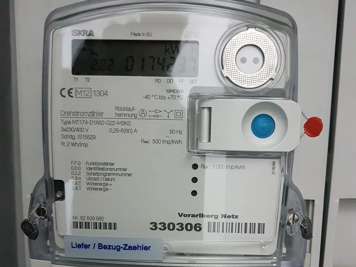 Grey three-phase electricity meter with a liquid crystal kWh display, an optical port and a label reading Liefer / Bezug-Zaehler, in a house's distribution cabinet
