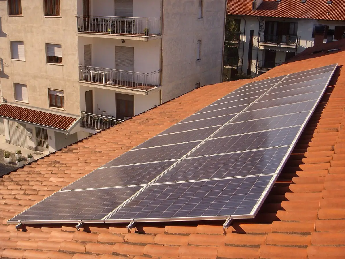 Rows of dark blue solar modules mounted on a terracotta tile roof beside apartment buildings in Italy