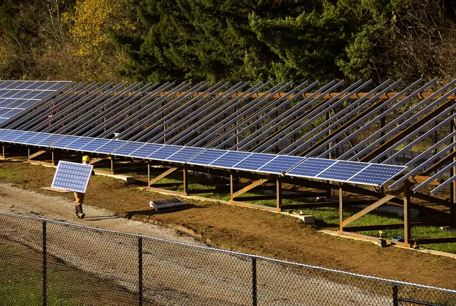 A worker in a hard hat carries one solar module across a gravel path beside rows of mounted modules